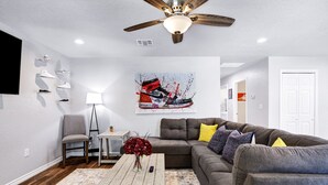 A living room with a ceiling fan, a gray couch with yellow and blue pillows, a wooden coffee table with a vase of red flowers, and a wall-mounted TV. - Rashali kicks theme house in SA (San Antonio)