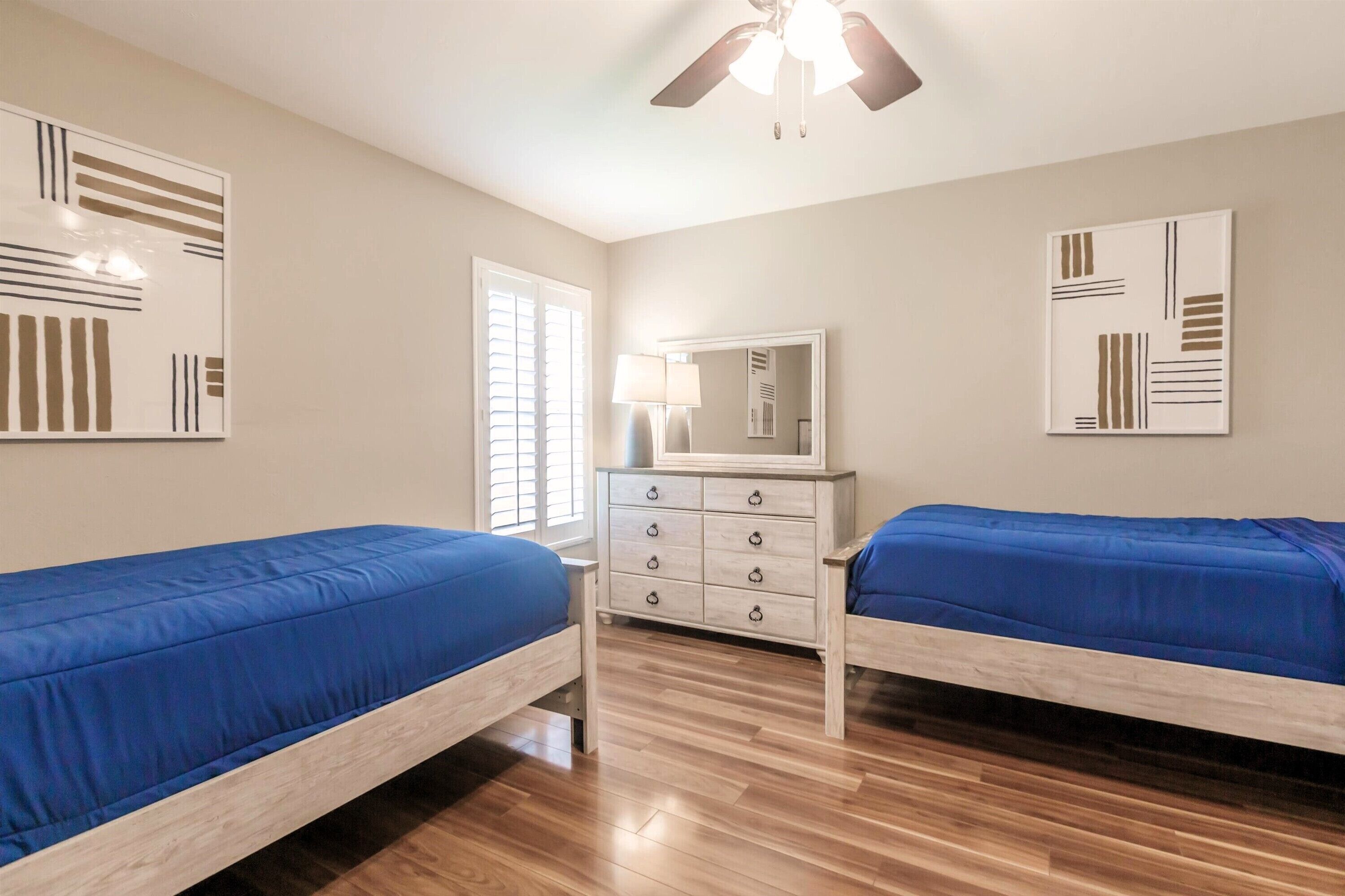 Une chambre avec deux lits simples, une commode, un ventilateur de plafond et des œuvres d’art accrochées aux murs.