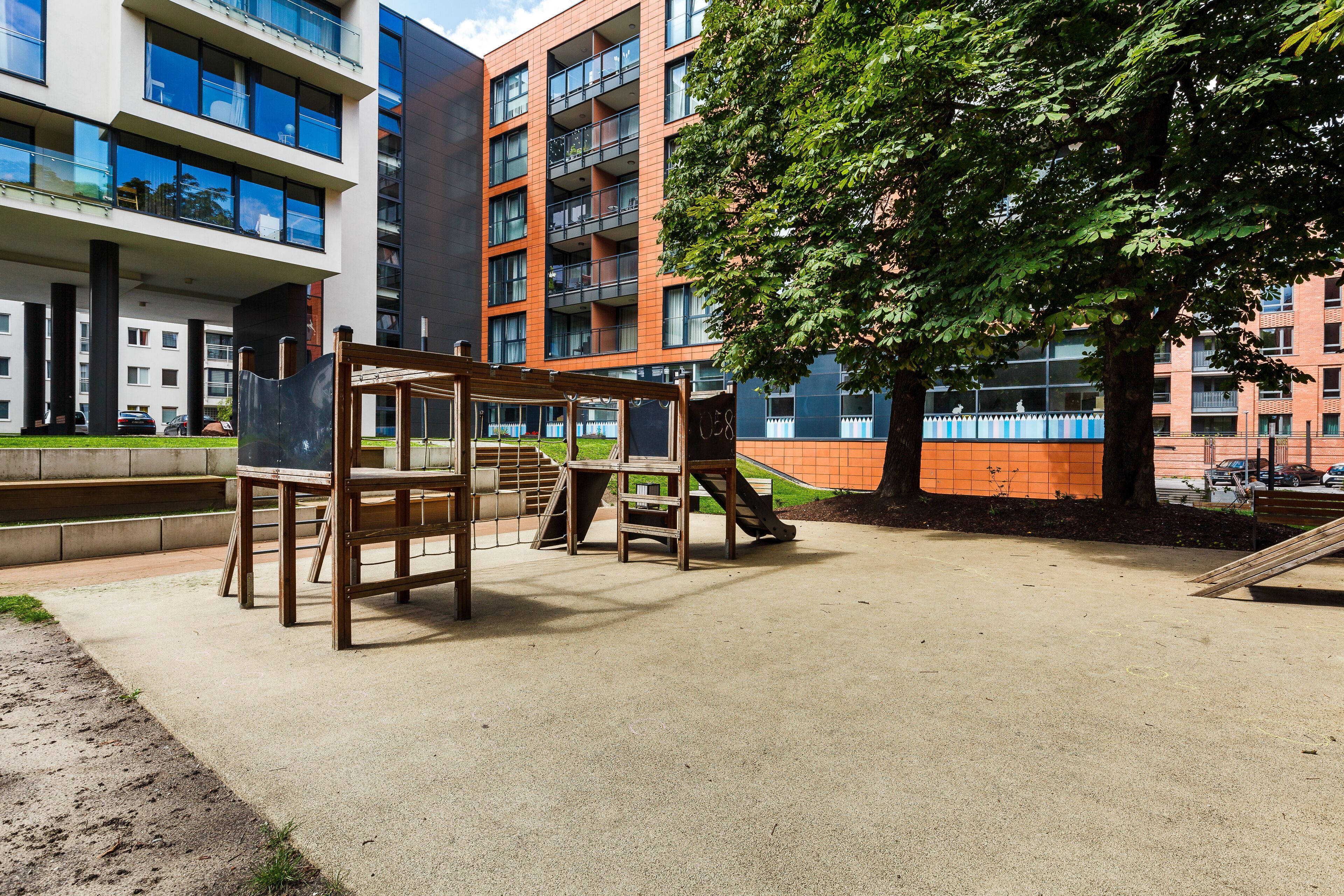 A playground with wooden structures and a concrete surface, surrounded by modern buildings with balconies and large windows.