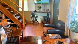 A living room with a wooden staircase, a leather sofa, a dining area with a bar, and a large TV displaying a scenic view. - NorthFork Lodge Cabin #2 "The Pine" (West)