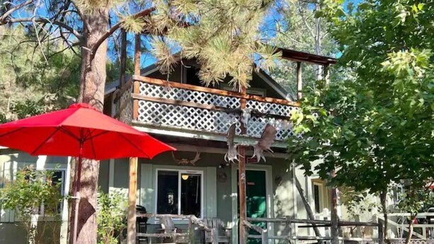 A rustic cabin with a balcony, outdoor seating, and a red umbrella. - NorthFork Lodge Cabin #2 "The Pine" (West)