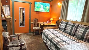 Ein gemütliches Zimmer mit Bett, Sessel, Tisch und Fernseher an der Wand.