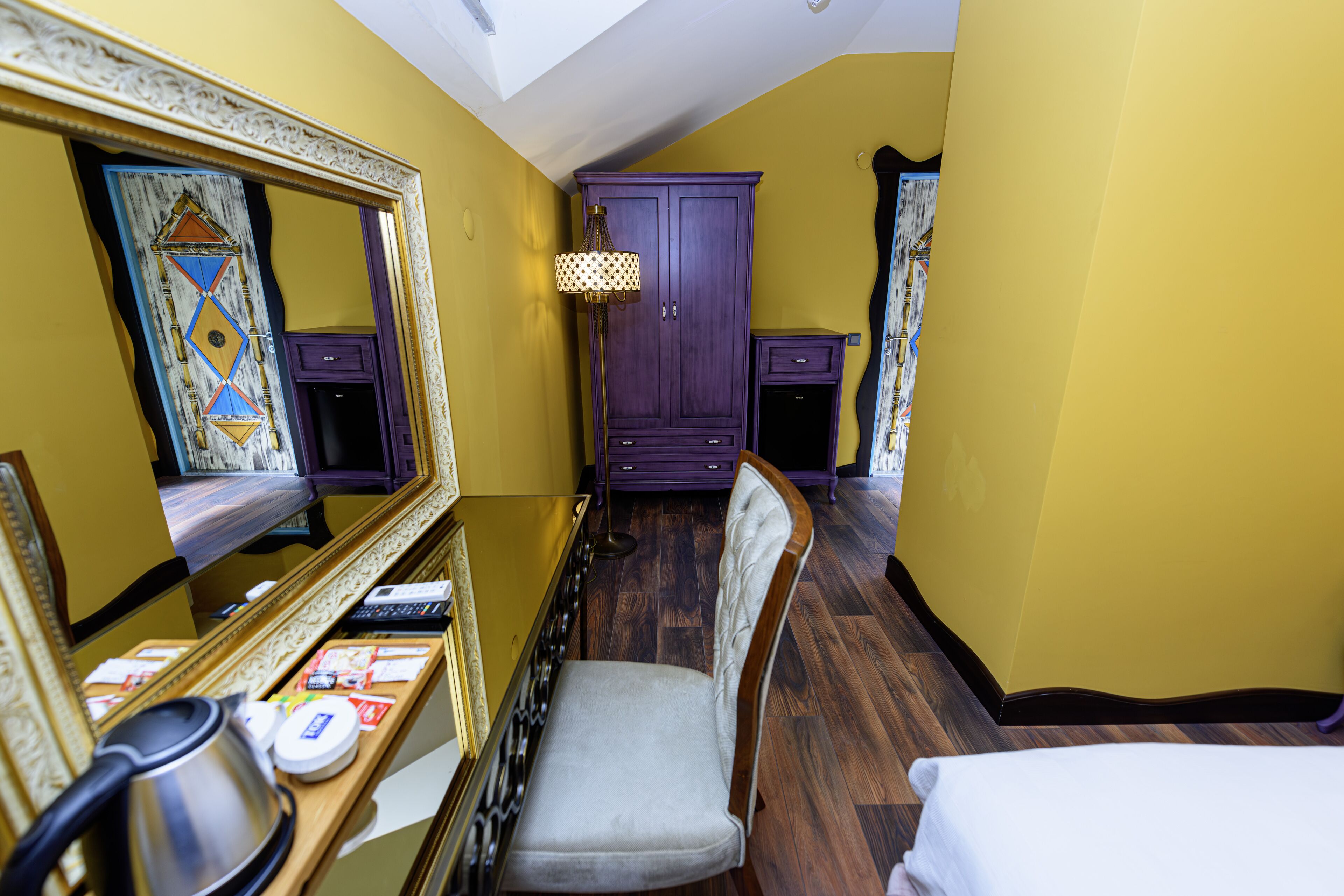 Una habitación de hotel con suelo de madera, un armario púrpura, un espejo, un escritorio con hervidor y una cama.