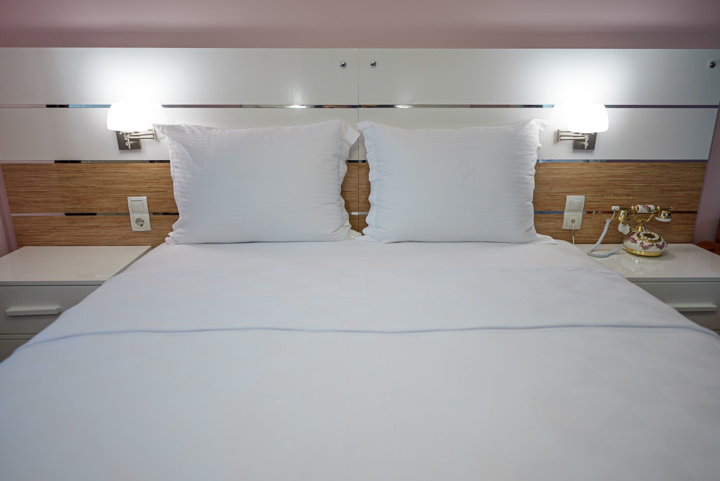 Quarto de hotel com uma cama grande, duas mesas de cabeceira, um painel de madeira na cabeceira e duas luminárias fixadas na parede.