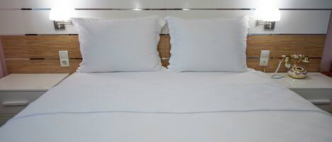 Quarto de hotel com uma cama grande, duas mesas de cabeceira, um painel de madeira na cabeceira e duas luminárias fixadas na parede.