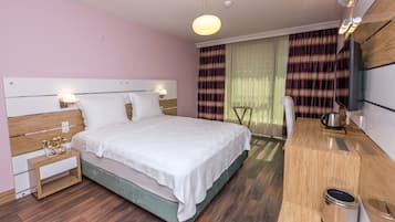 A modern hotel room with a large bed, bedside tables, a TV, and a wooden desk with a kettle.