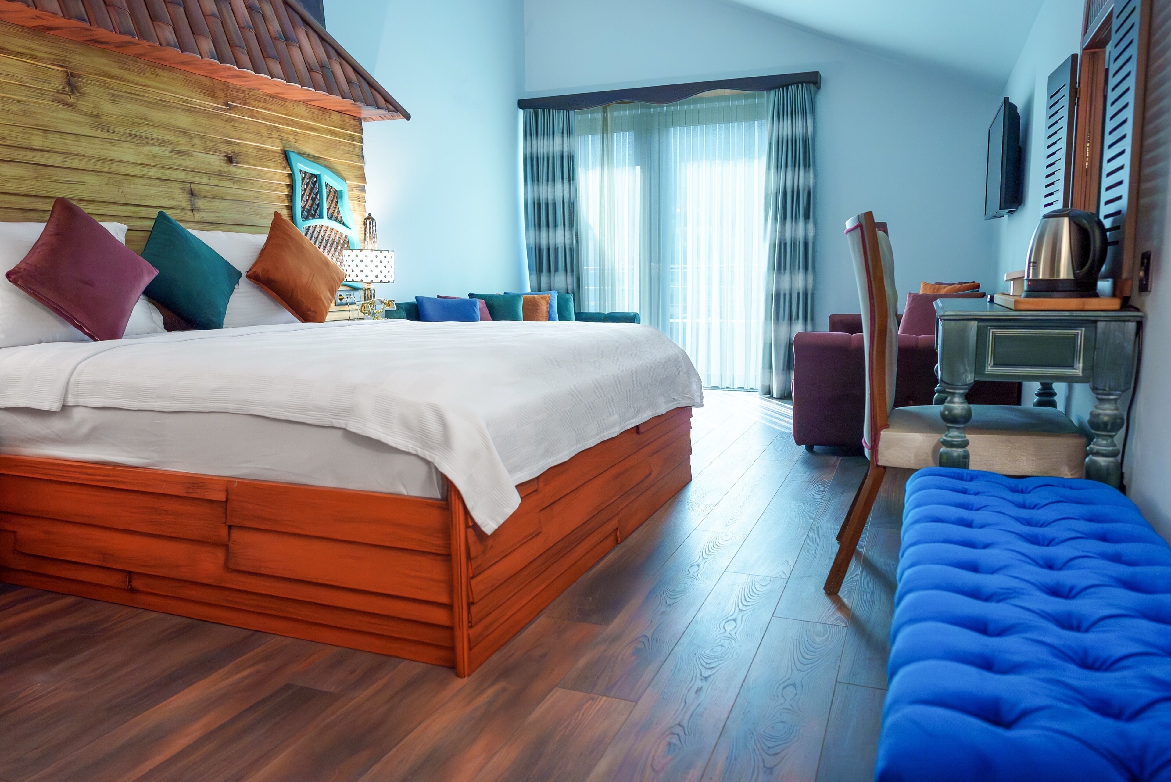 A bedroom with a wooden headboard, a large bed with multiple pillows, a blue ottoman, a wooden dresser, and a painting on the wall.