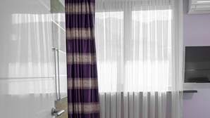 A modern kitchen with white cabinets, a wooden floor, a window with sheer white curtains and a purple striped curtain, and a built-in air conditioning unit. - TOK EPİK HOTEL (Bursa)
