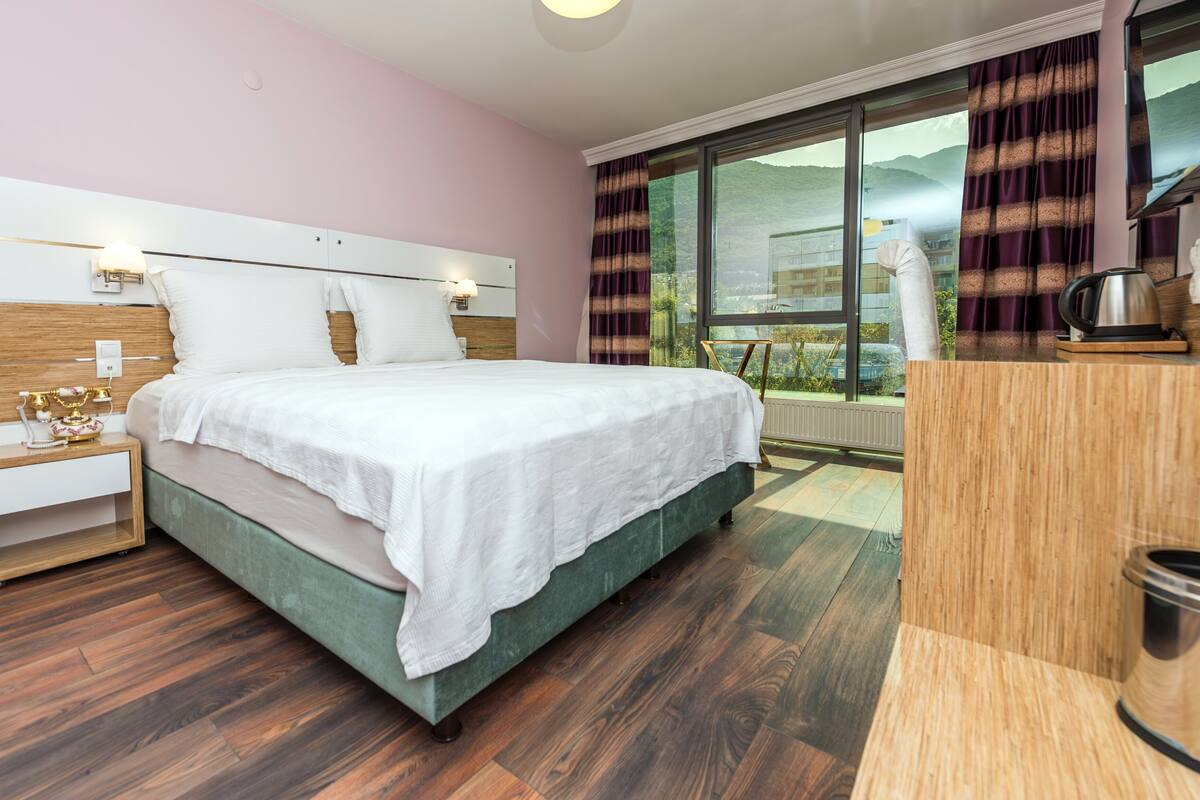 Un dormitorio con una cama grande, piso de madera, una mesita de noche con lámpara y vistas al exterior a través de una ventana.