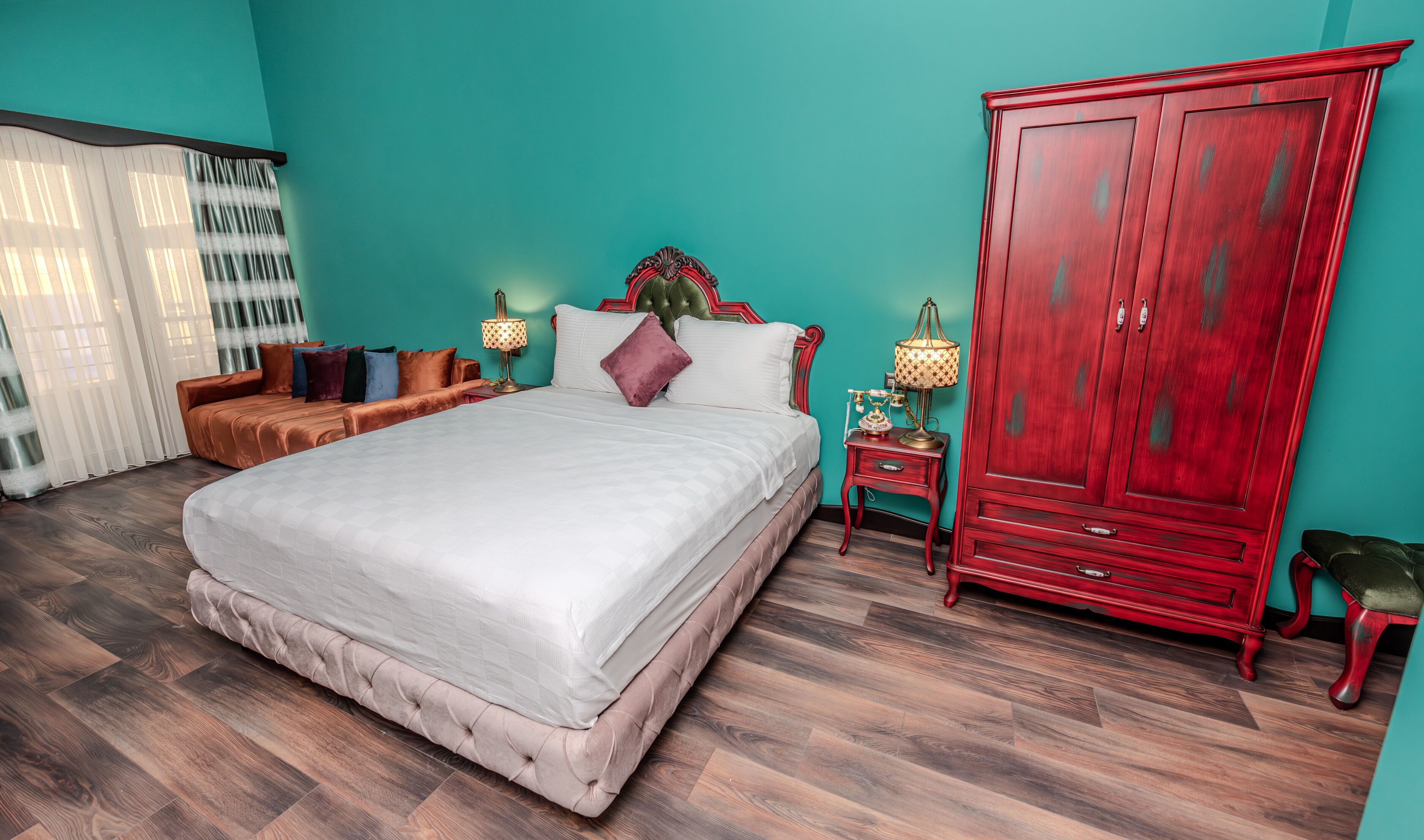 A bedroom with a large bed, a red wardrobe, a nightstand with lamps, and a window with sheer curtains.