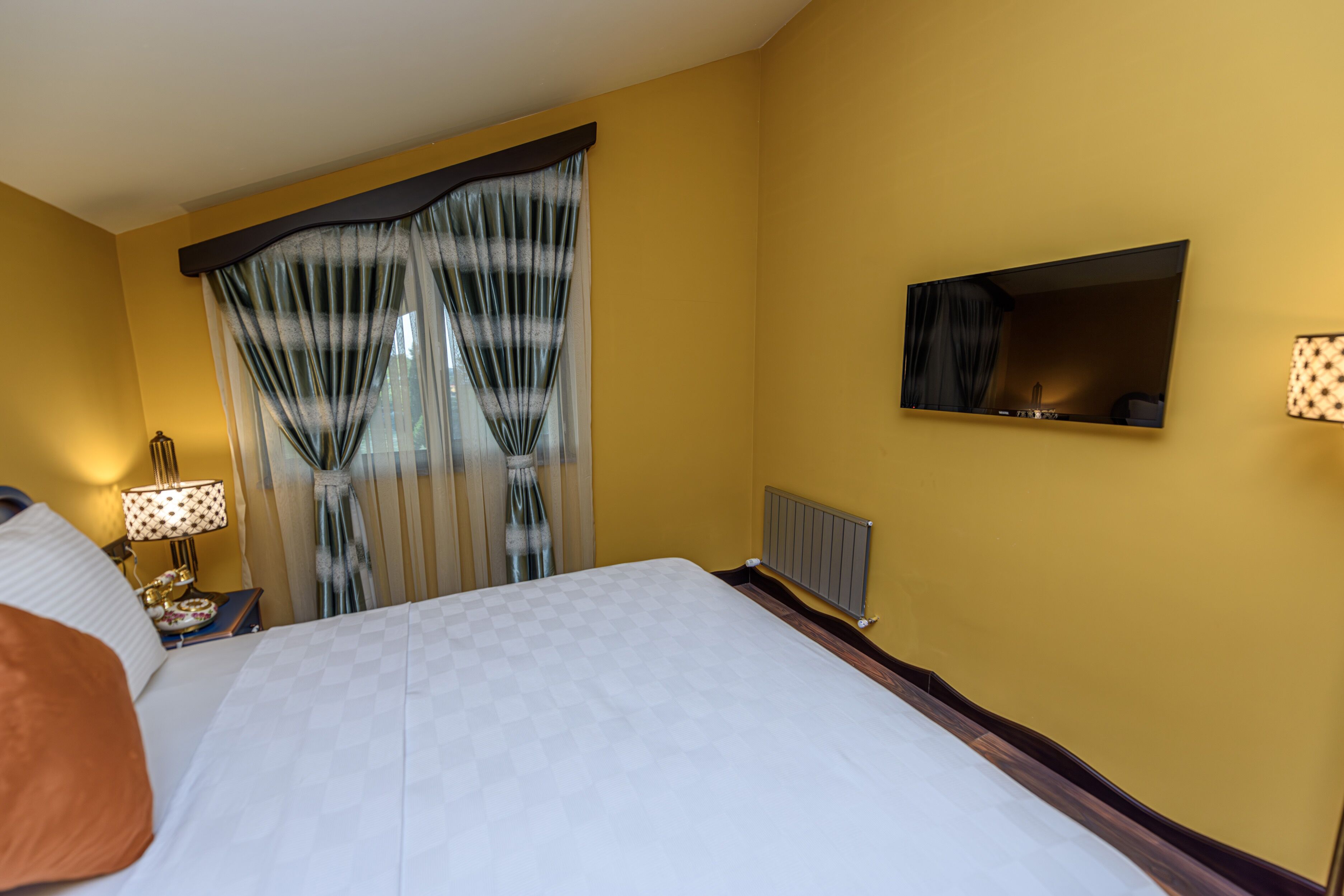 A hotel room with a large bed, a nightstand with a lamp, a mirror on the wall, and sheer curtains on the window.
