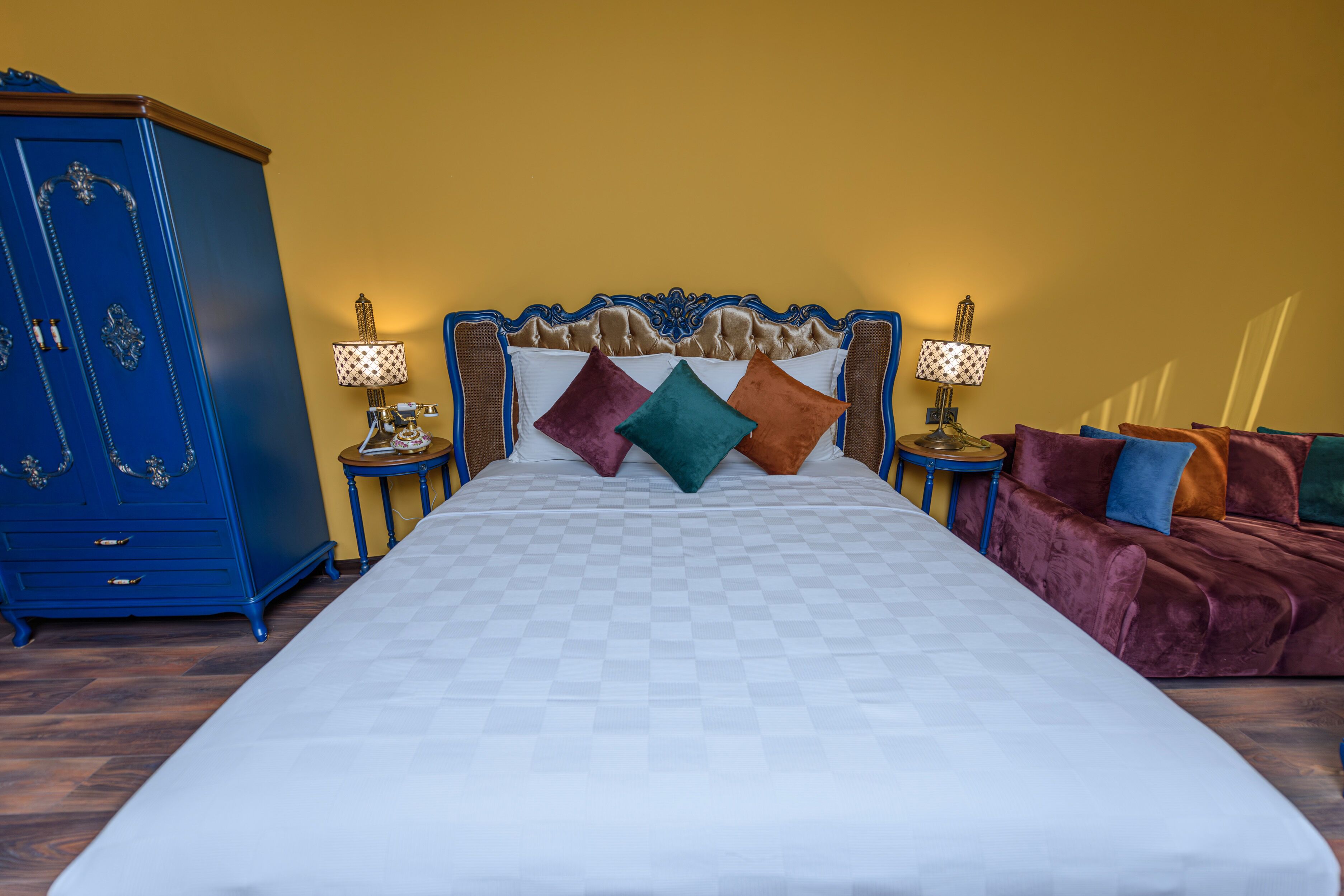 A neatly made bed with a checkered bedspread, a headboard with decorative carvings, and a blue wardrobe to the left.