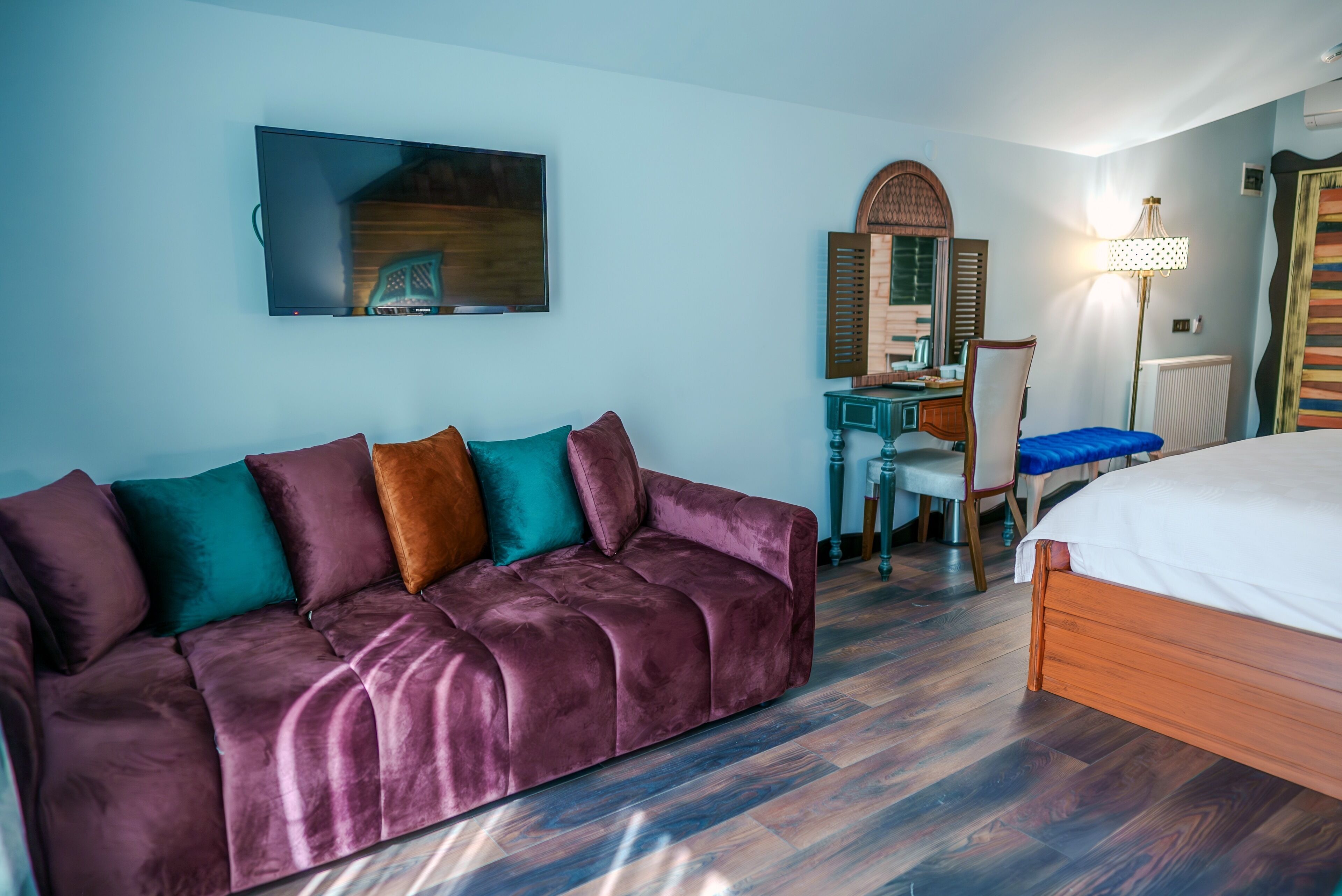 A room with a large purple sofa, a wooden bed, a desk with a chair, a mirror, and a television mounted on the wall.