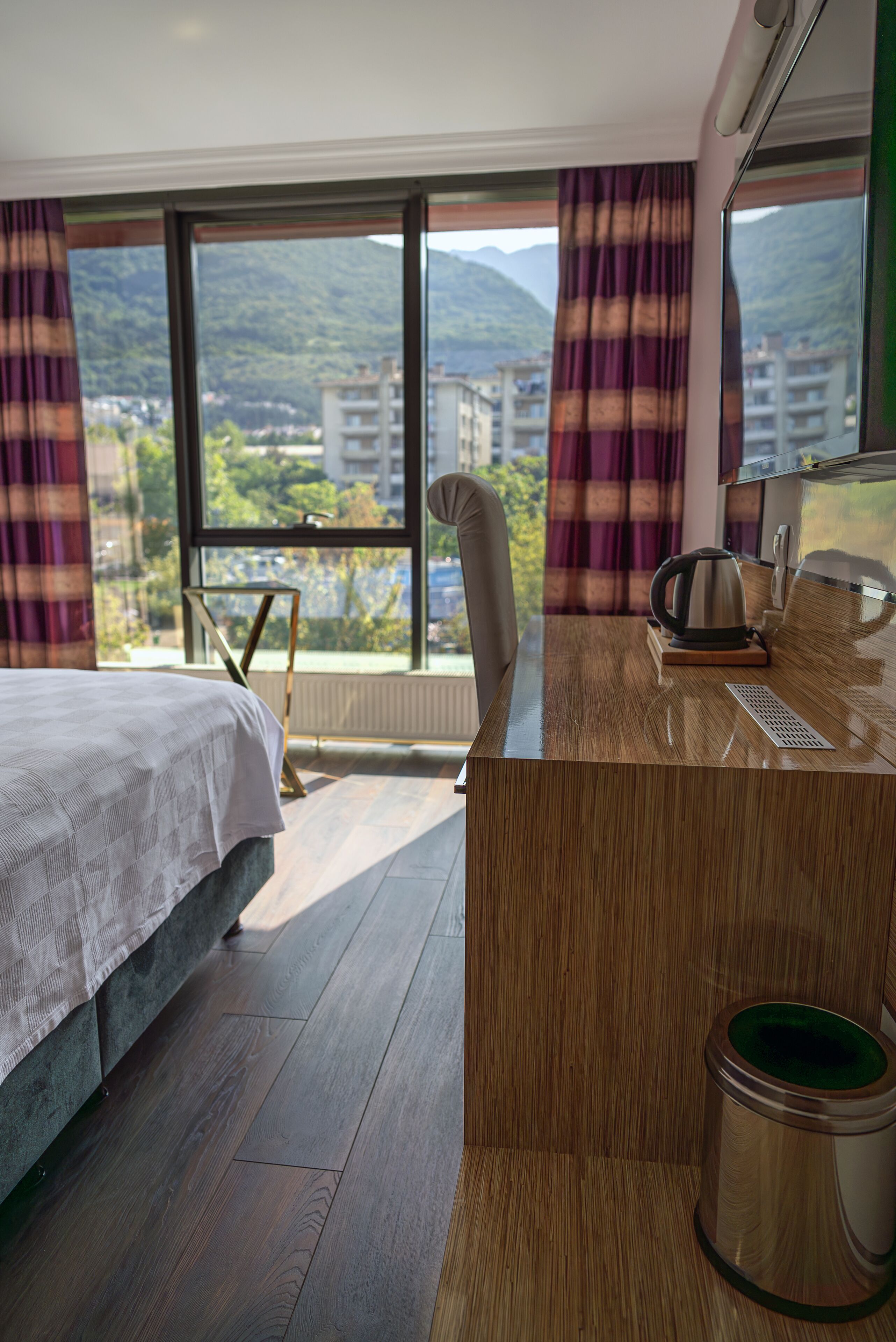 A hotel room with a bed, a desk with a kettle, a trash bin, and large windows offering a mountain view.
