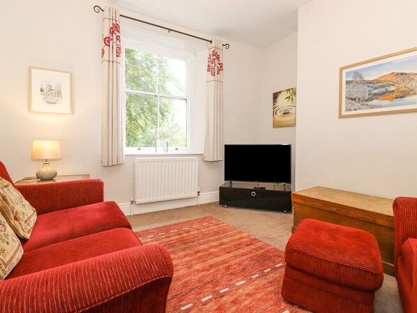 A room with a red sofa set, a flat-screen TV, a wooden chest of drawers, and a window with curtains. - Riverbank (Ambleside)