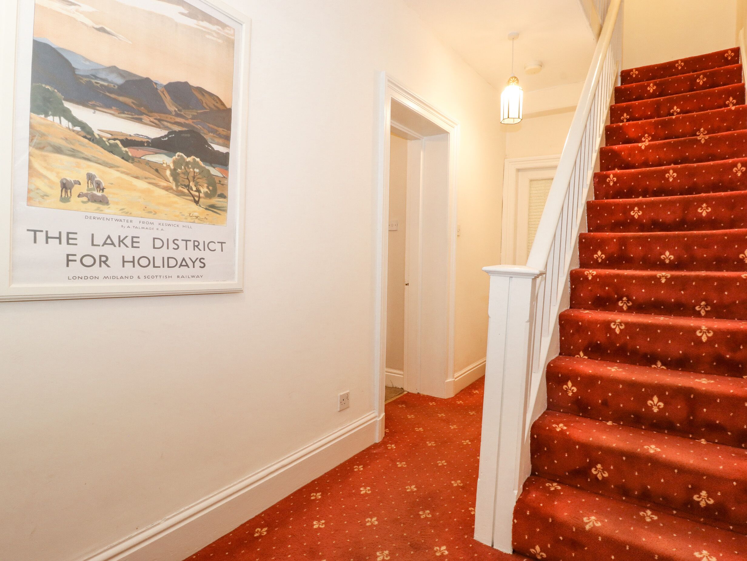 A staircase with red carpet and white balusters, leading to a room with white walls and a framed poster of a lake district.
