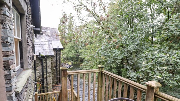 A wooden deck with a metal railing overlooking a river, surrounded by lush greenery. - Riverbank (Ambleside)