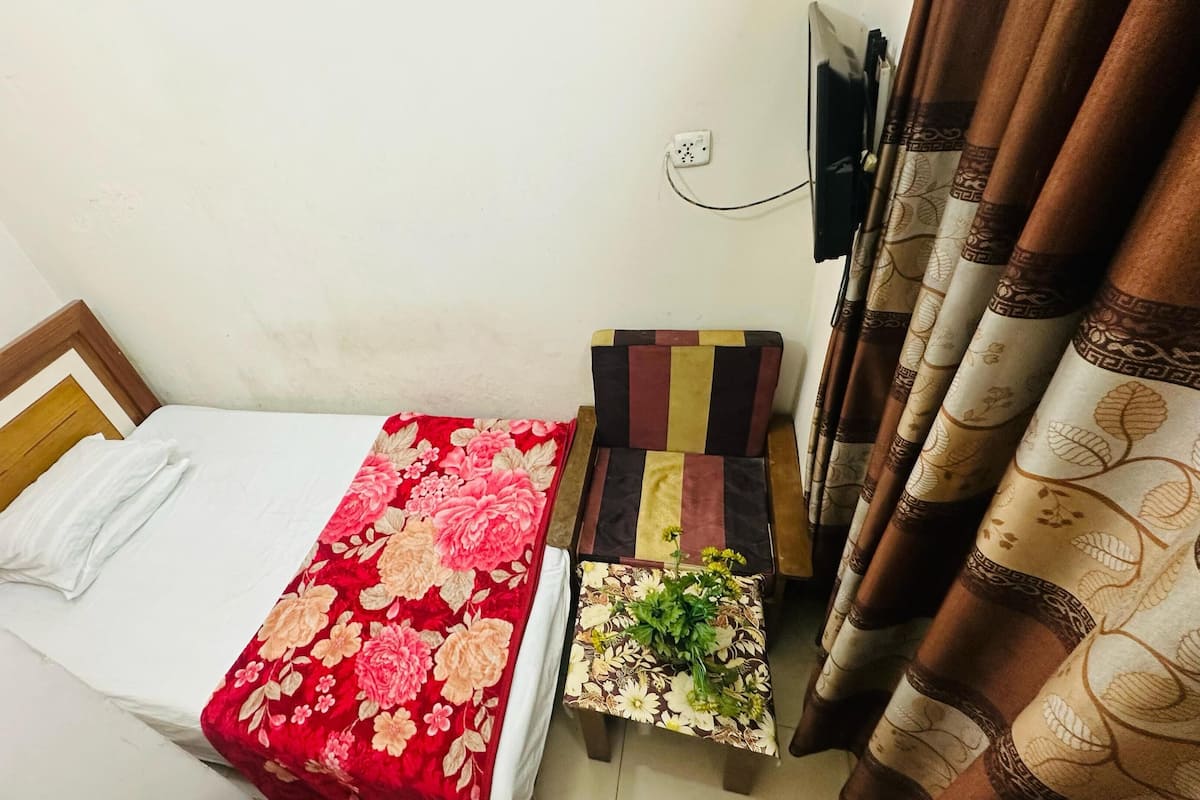 Une petite pièce, de taille modeste, comprenant un lit, une petite table recouverte d’une nappe à motifs floraux, un téléviseur fixé au mur et des rideaux ornés de motifs de feuilles.