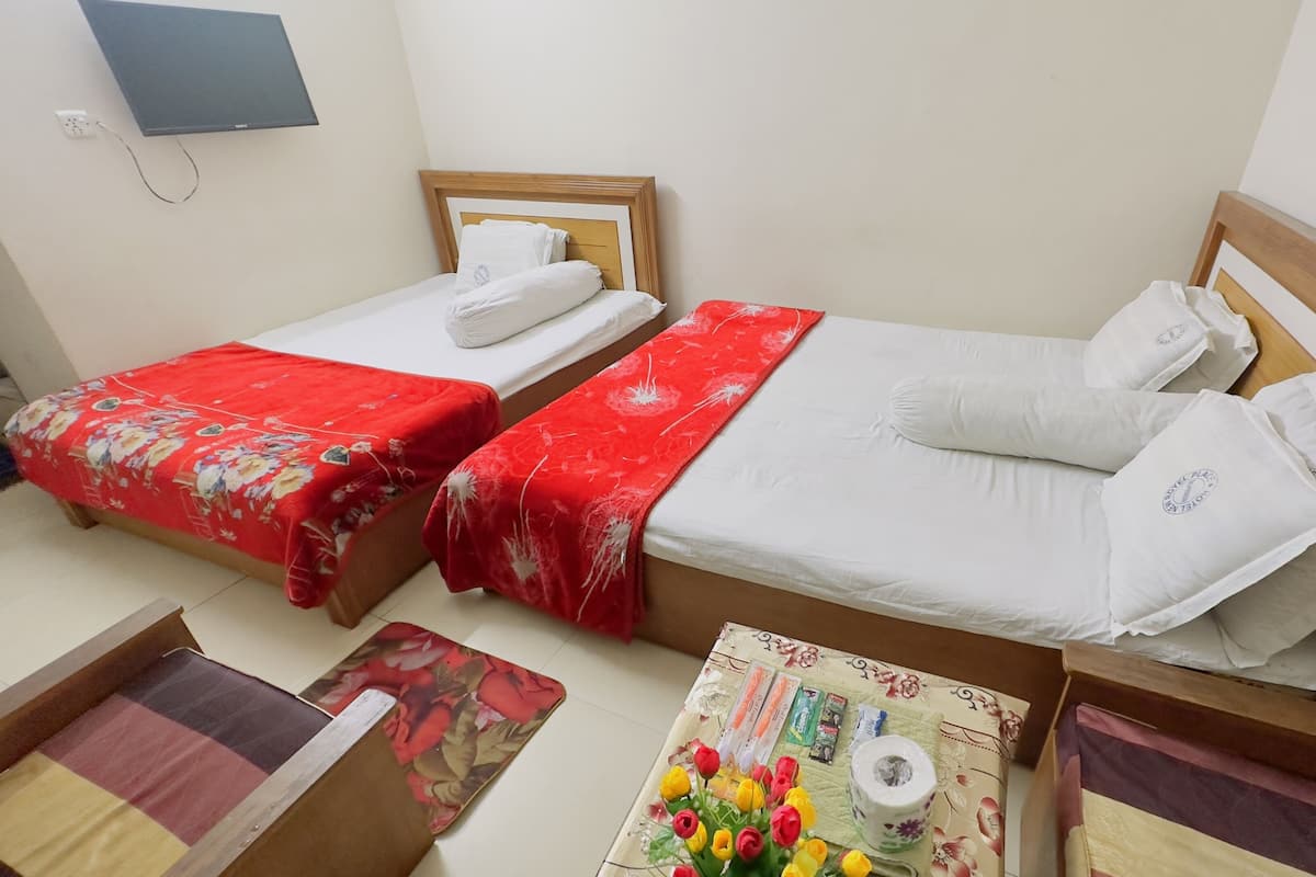 Une chambre d’hôtel avec deux lits simples, chacun doté d’une tête de lit en bois, d’une couvre-lit à motifs floraux rouges et de draps blancs. On y trouve une petite table recouverte d’un chemin de table à motifs floraux, un vase avec des fleurs artificielles et quelques articles de toilette.