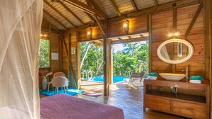 A wooden cabin with a bed, a bathroom with a sink and mirror, and a view of a swimming pool.