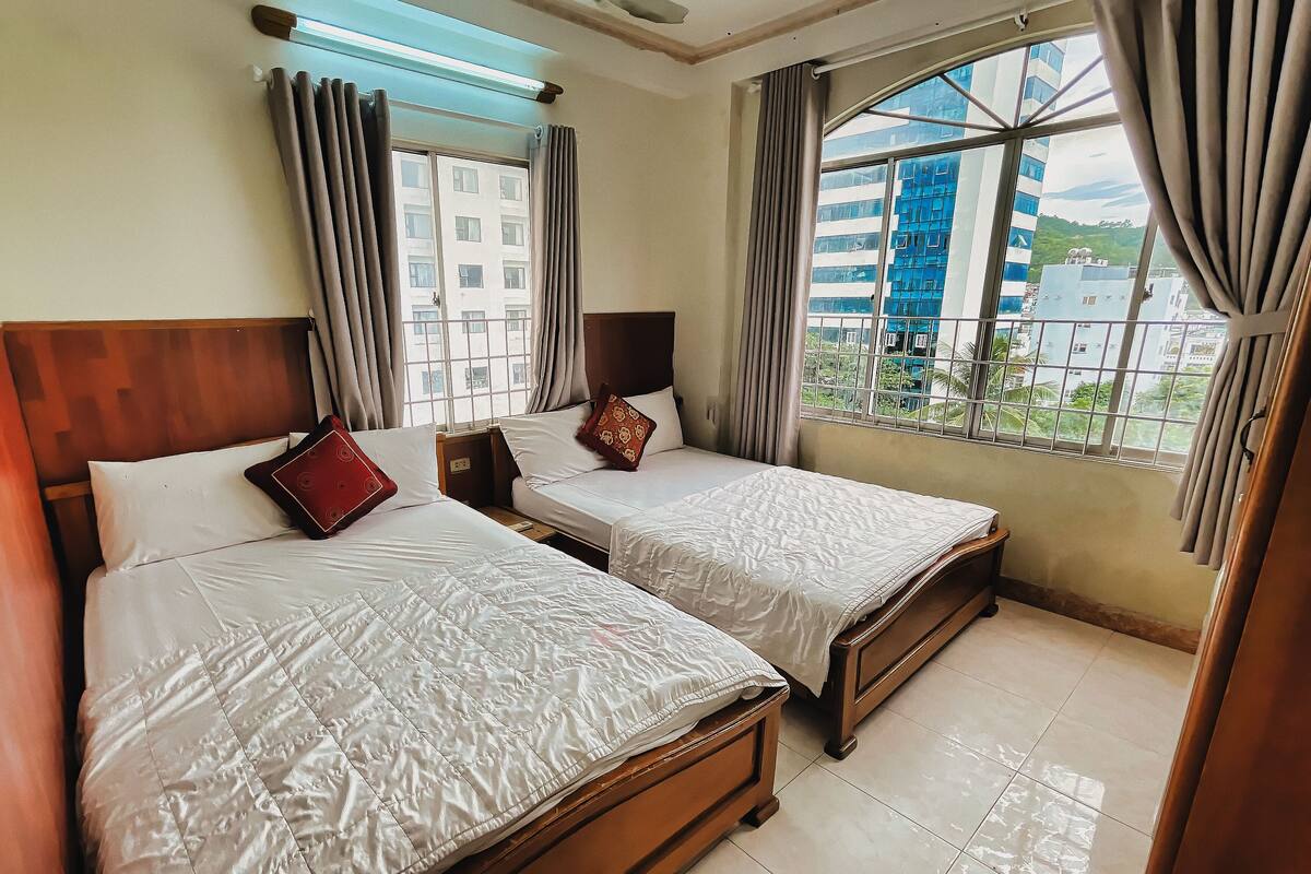 A hotel room with two beds, a large window with curtains, and a view of buildings outside.