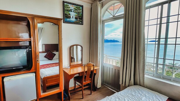 A hotel room with a bed, a desk with a chair, a microwave, a mirror, and a large window with a view of the sea. - Quang Vinh 2 Hotel (Bac Nha Trang)