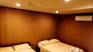 A hotel room with two single beds, each with a white bedspread and pillows, and a towel on the bed. There is a ceiling light and an air conditioning unit on the wall.