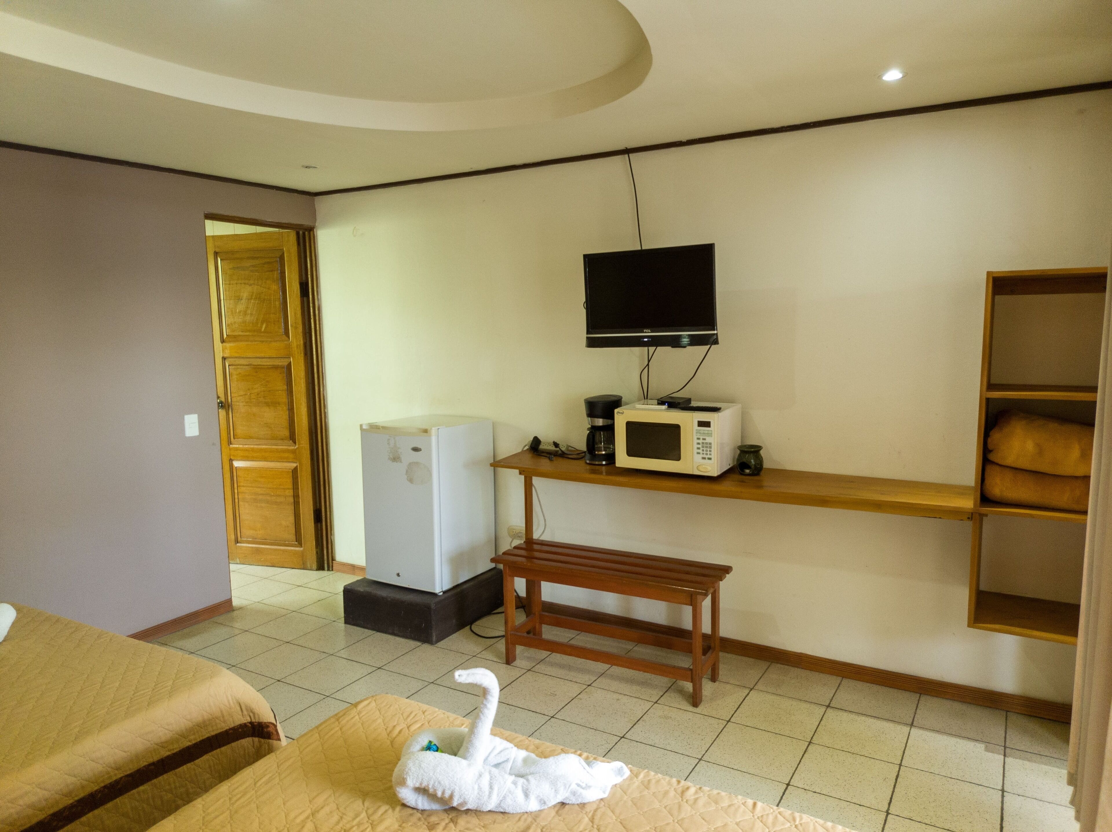 A hotel room with a bed, a small wooden table, a microwave, a TV, and a refrigerator.
