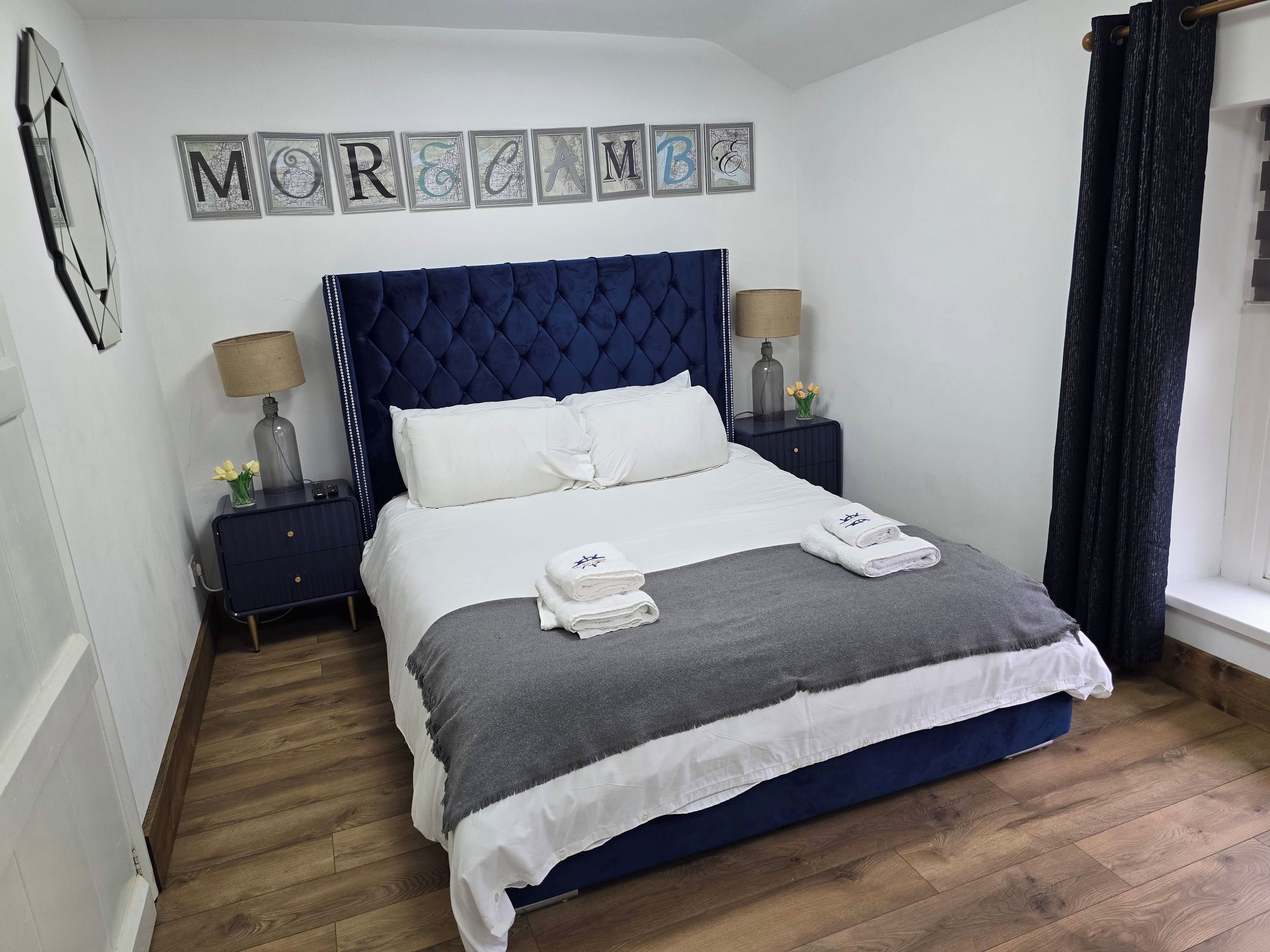 Un lit bien fait, avec une tête de lit bleue, du linge de lit blanc et deux serviettes pliées. Il y a deux tables de chevet avec des lampes et un petit vase de fleurs. Le mur au-dessus du lit est décoré d’œuvres d’art encadrées, représentant des lettres.