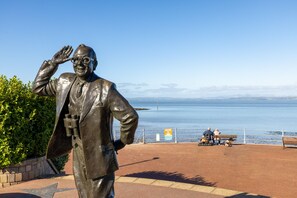 Una estatua de un hombre trajeado con el brazo levantado, contemplando un paseo marítimo con bancos y un cielo despejado.