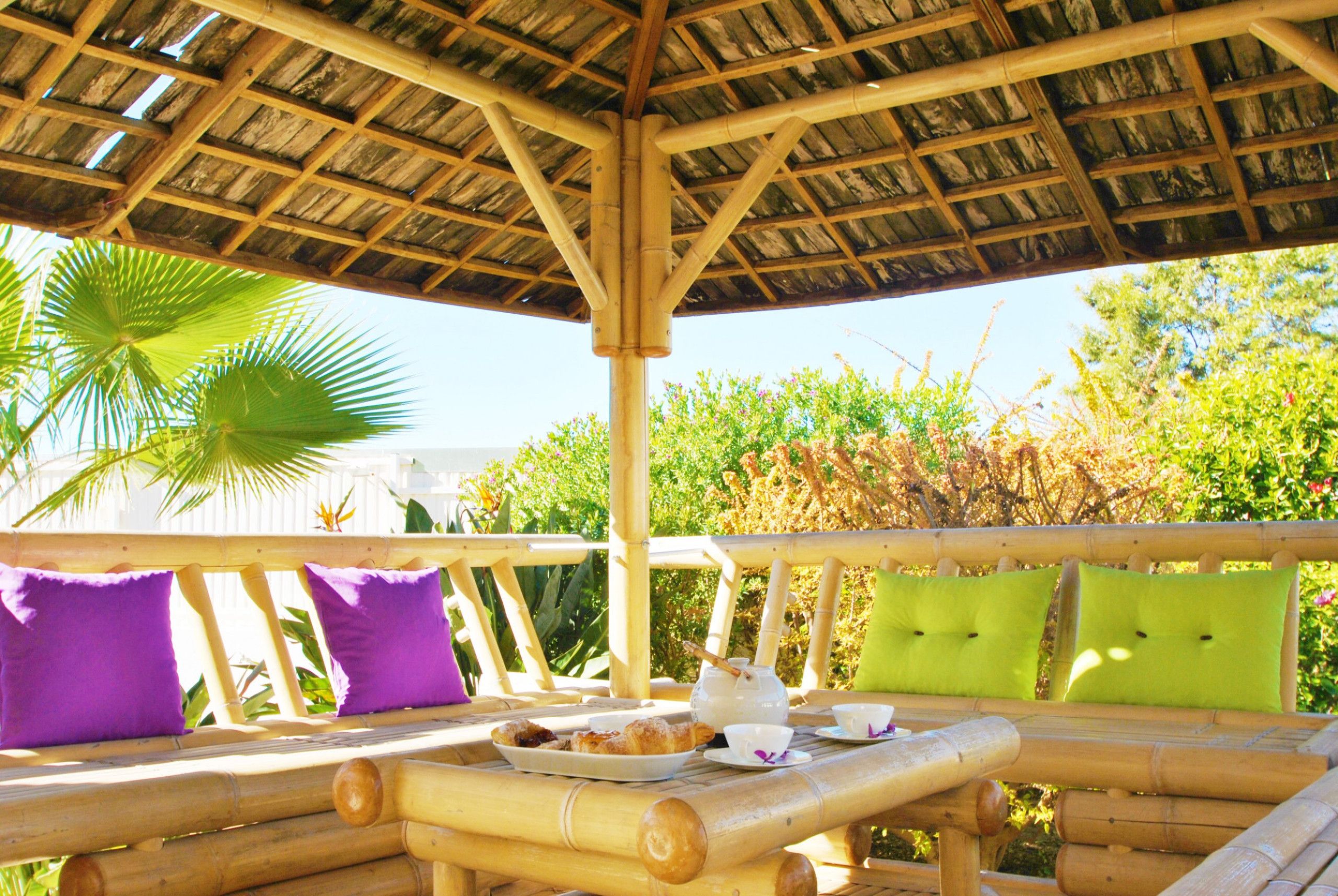 An outdoor seating area with bamboo furniture and cushions, a table with a teapot and cups, and a thatched roof structure.