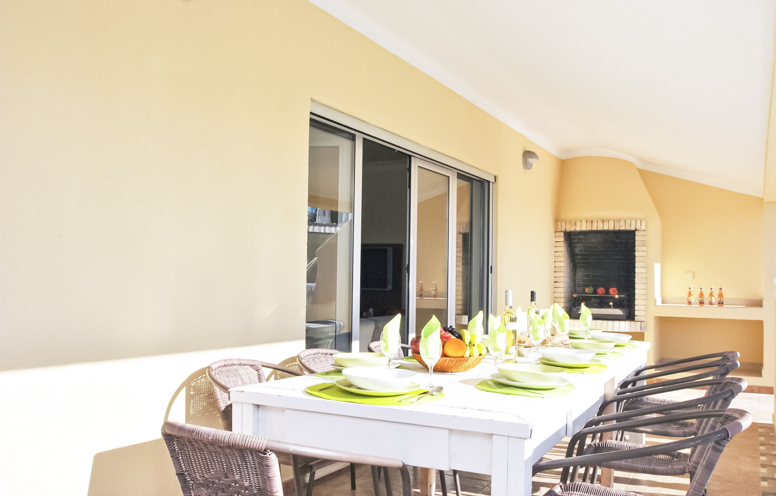 A dining area with a white table set for six, surrounded by wicker chairs, a glass door, and a brick fireplace.