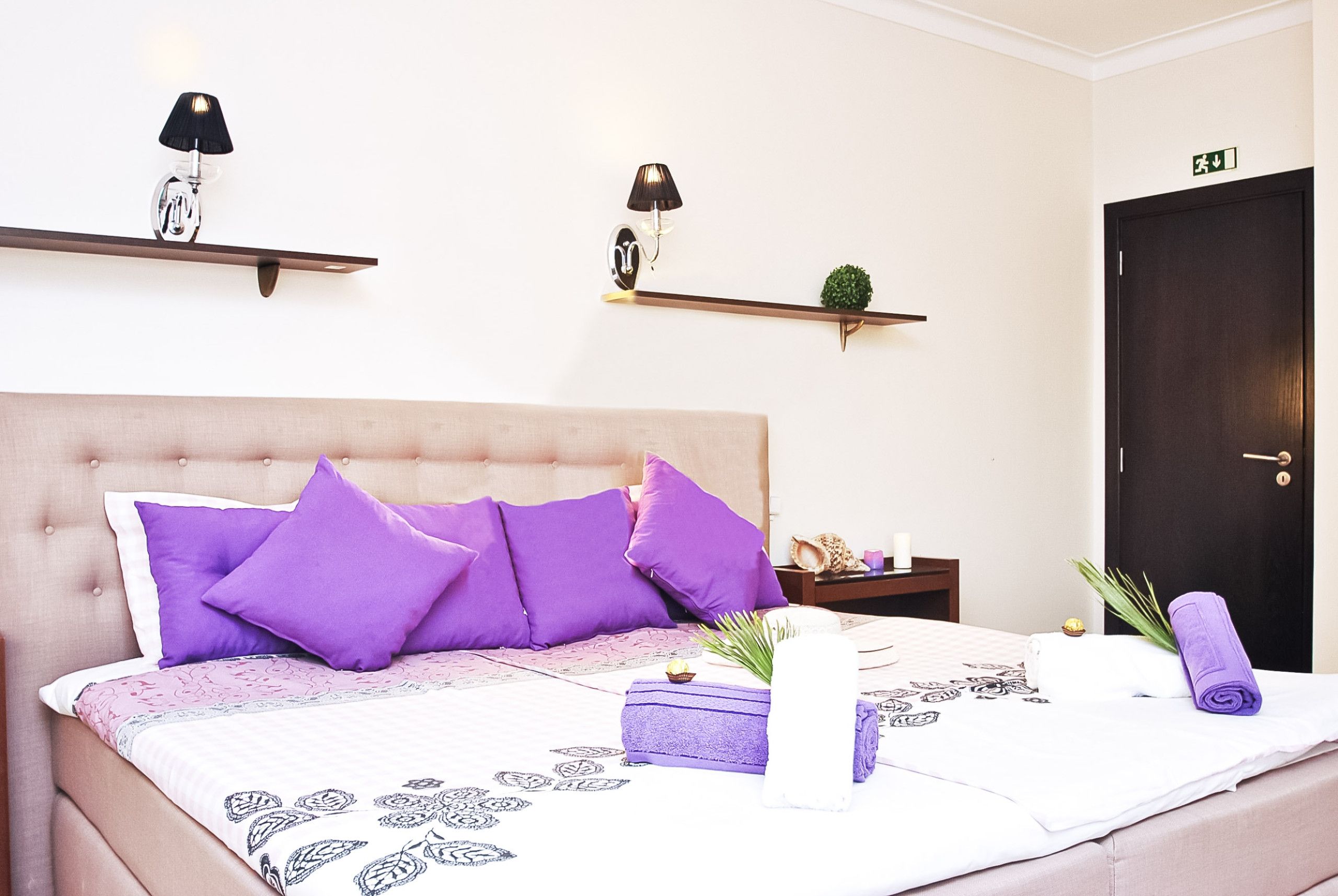 Una cama bien hecha con cabecera, varios almohadones, una mesita de noche con lámpara, una planta pequeña y una puerta con letrero de salida.