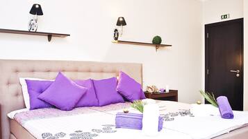 Una cama bien hecha con cabecera, varios almohadones, una mesita de noche con lámpara, una planta pequeña y una puerta con letrero de salida.