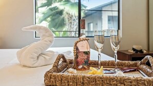 A neatly arranged breakfast tray with a bottle of Beach rum, two glasses, and a white towel on a bed with a view of palm trees through a window. - Top Floor updated condo, oceanview, AC, walk to shops, dining, and beach (Kihei)