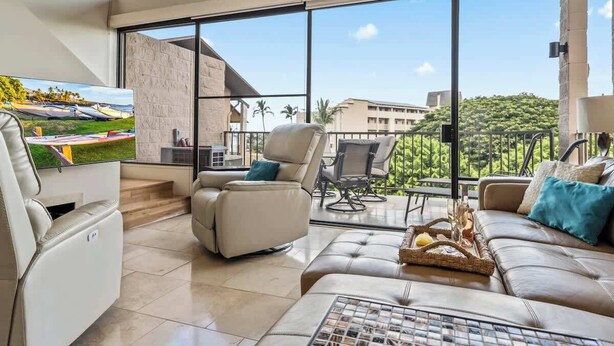 A modern living room with a large window, a sofa, a recliner, and a flat-screen TV. - Top Floor updated condo, oceanview, AC, walk to shops, dining, and beach (Kihei)