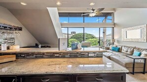 A modern kitchen with a large island, a dining area with a sofa, and a view of greenery outside. - Top Floor updated condo, oceanview, AC, walk to shops, dining, and beach (Kihei)
