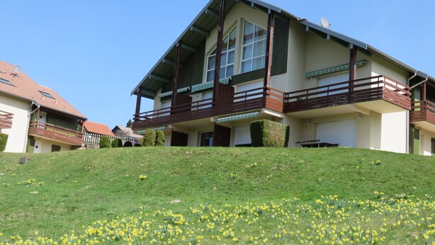A two-story building with a balcony on the upper floor, surrounded by a grassy area with yellow flowers. - Appartement Vacances (Gérardmer)