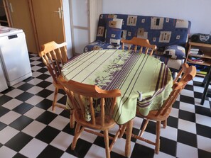 Uma sala de jantar com piso xadrez, uma mesa redonda coberta por toalha florida e cadeiras de madeira.
