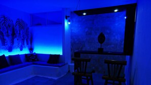 A room with a blue-lit wall, a sofa with cushions, two wooden chairs, and a small table with a decorative item. - 1 Deluxe Lifestyle (Sosúa)