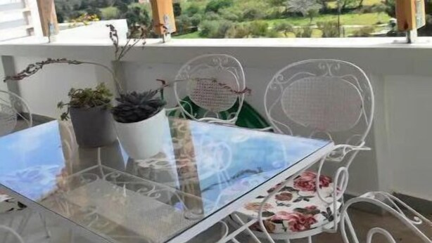 Un balcon avec une table en verre et des chaises en métal blanc, donnant sur un paysage verdoyant avec des collines.