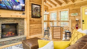 A cozy log cabin interior with a fireplace, wooden walls, a dining area, and a television mounted on the wall. - Harper’s Hideaway- Close to Downtown Gatlinburg, Hot Tub, Guest Favorite! (Gatlinburg)