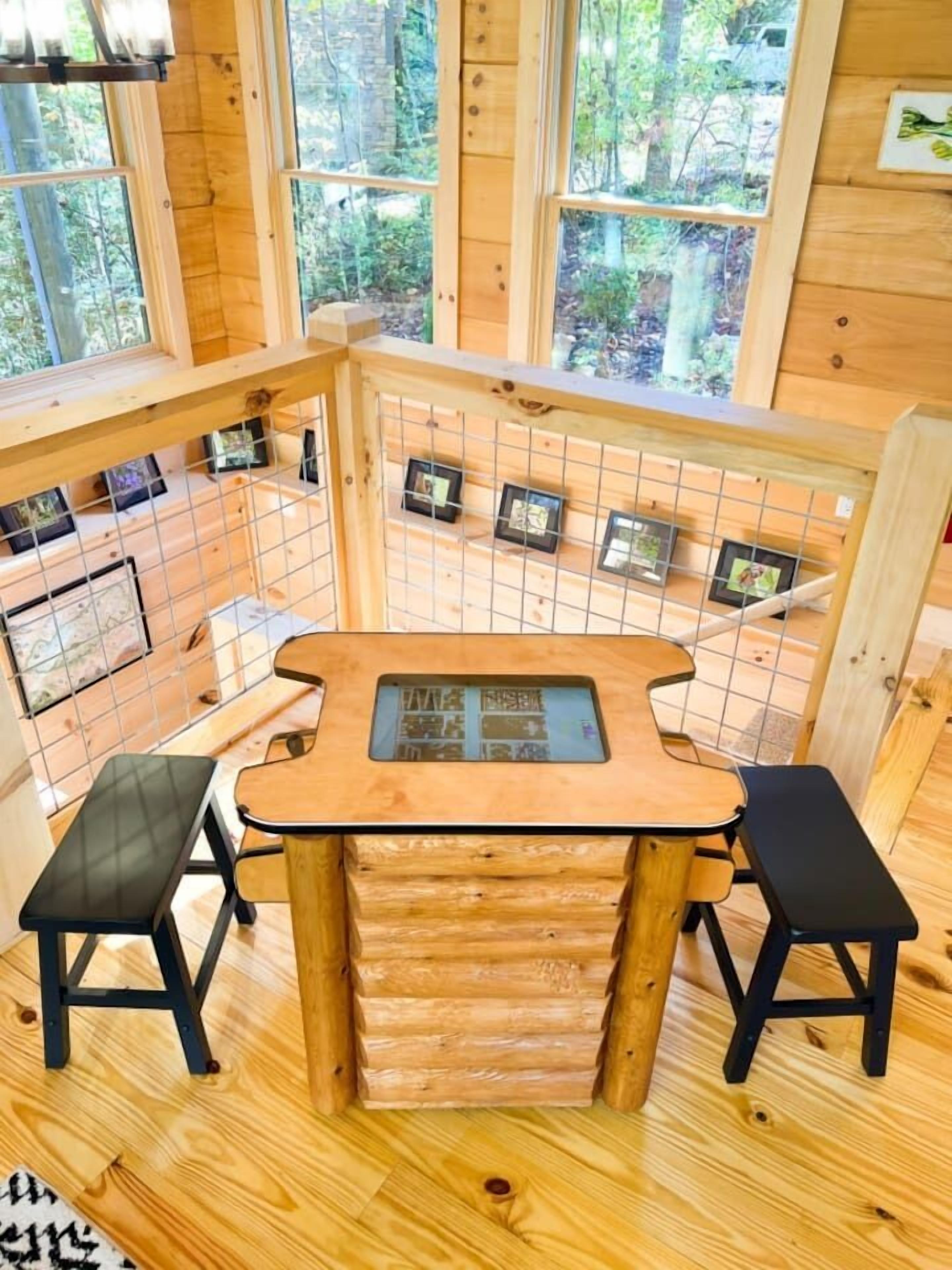 A wooden cabin with a central table and two benches, surrounded by large windows offering a forest view.