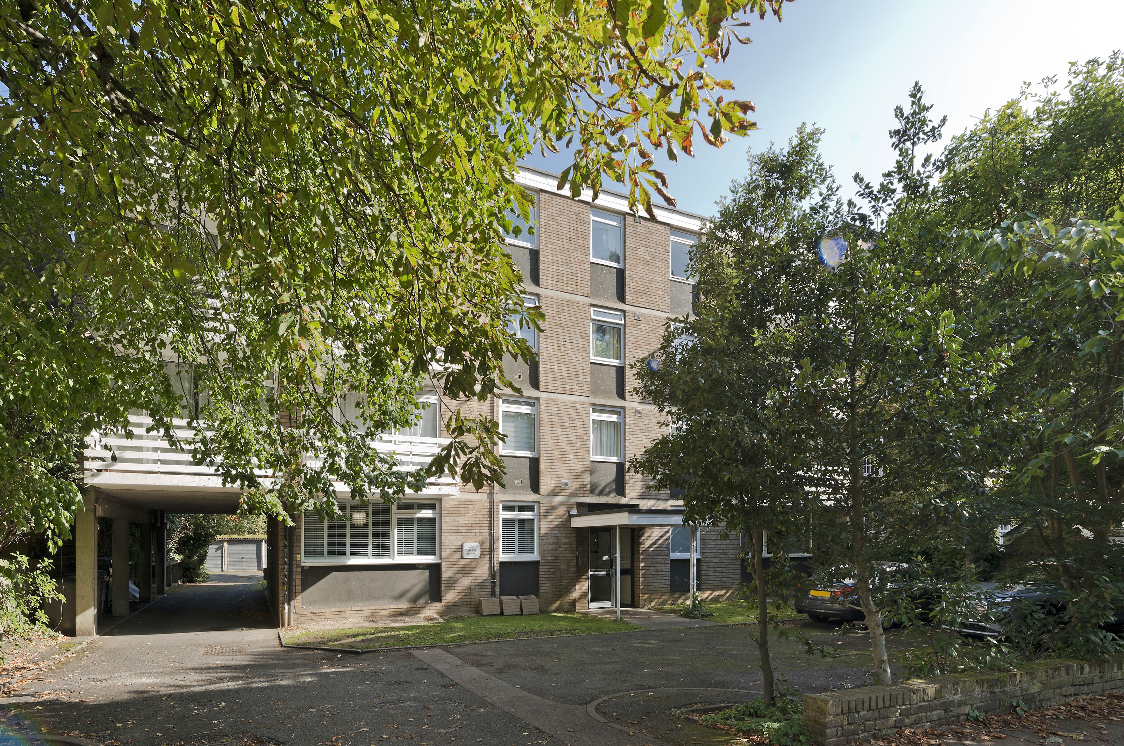 A multi-story residential building with a covered entrance, a small garden area, and parked cars.