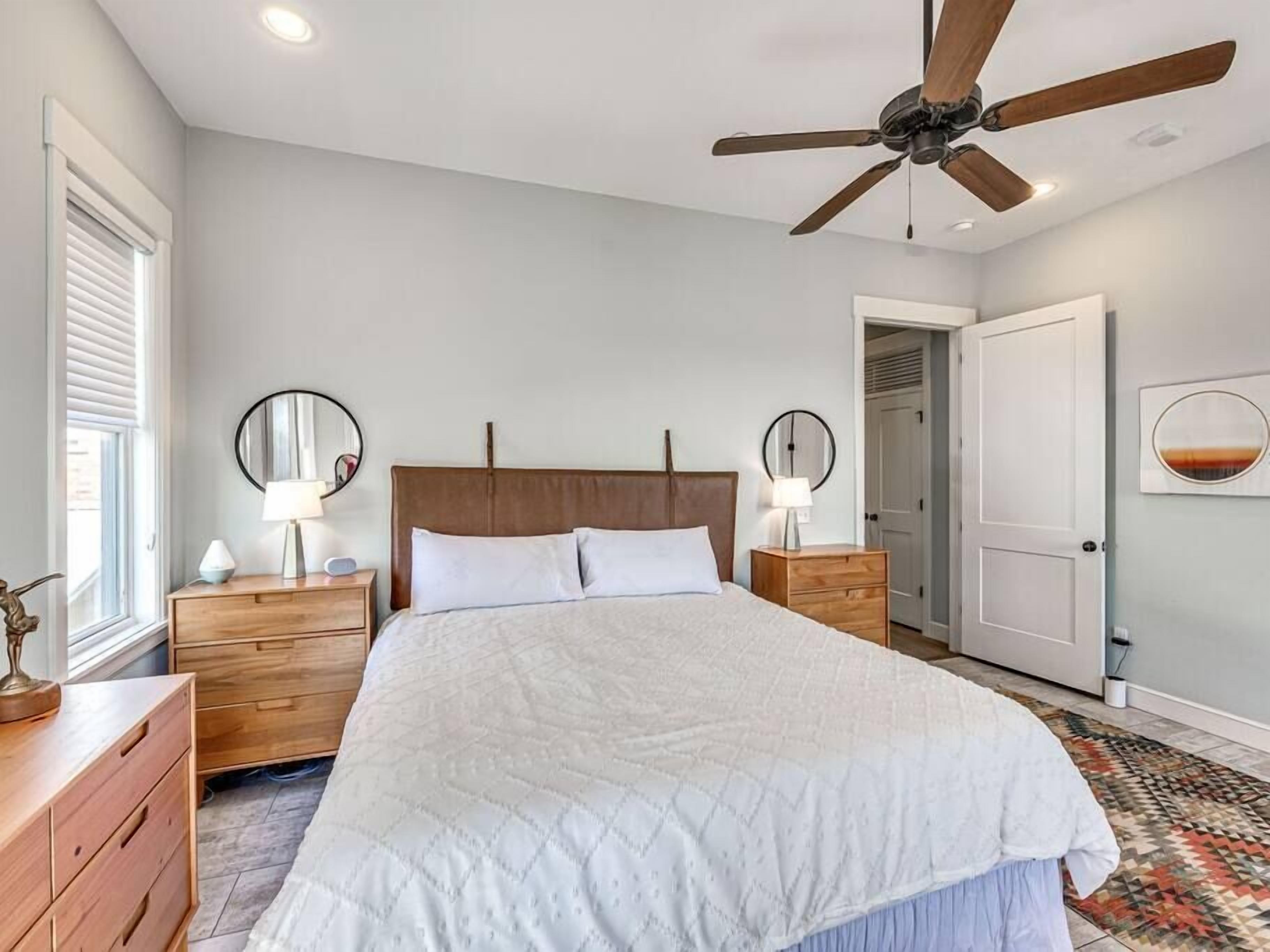 Une chambre à coucher comprenant un lit, des tables de chevet, un ventilateur de plafond, une porte et un miroir fixé au mur.