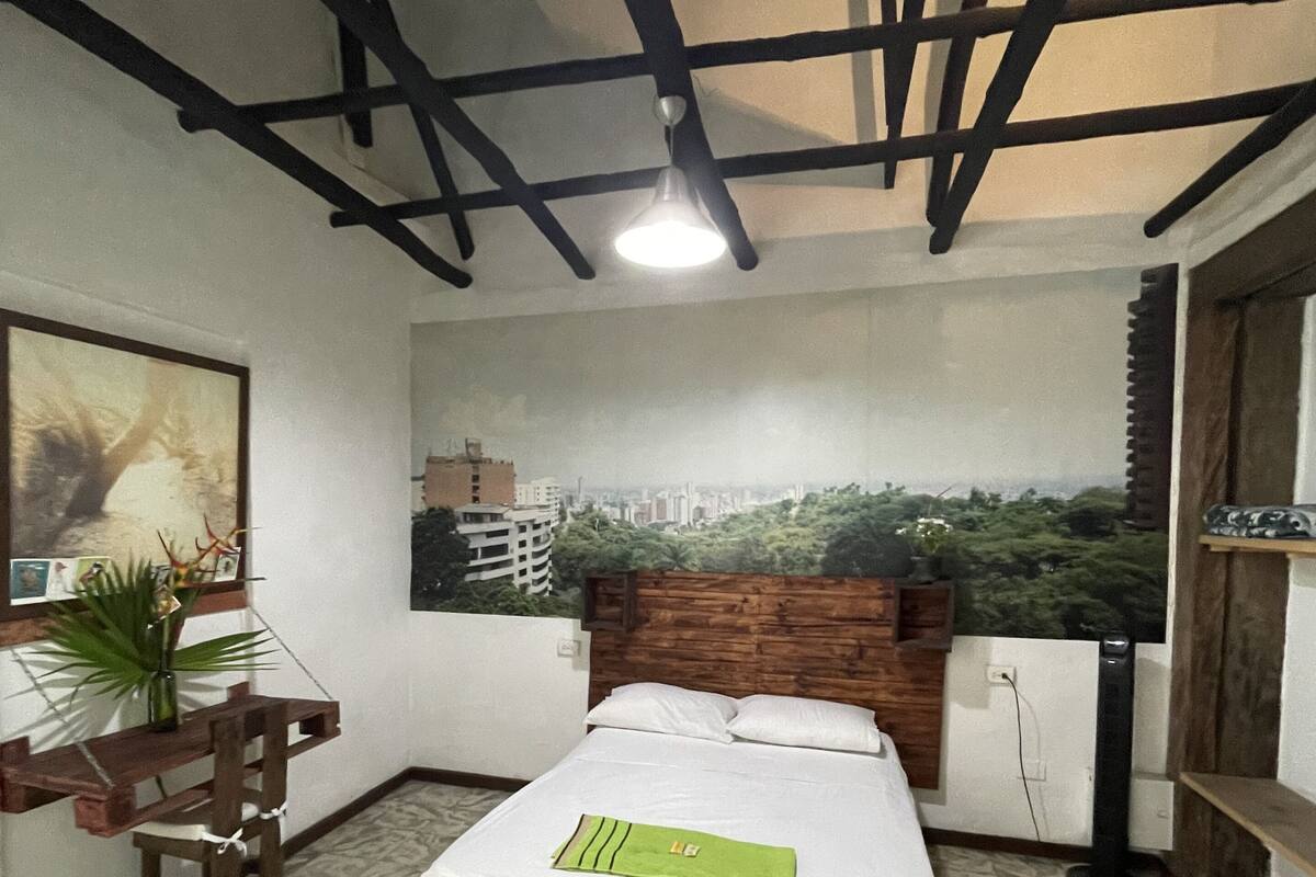 Une chambre à coucher avec un lit, un bureau et une chaise. Un grand tableau mural représentant un paysage urbain orne le mur situé derrière le lit.