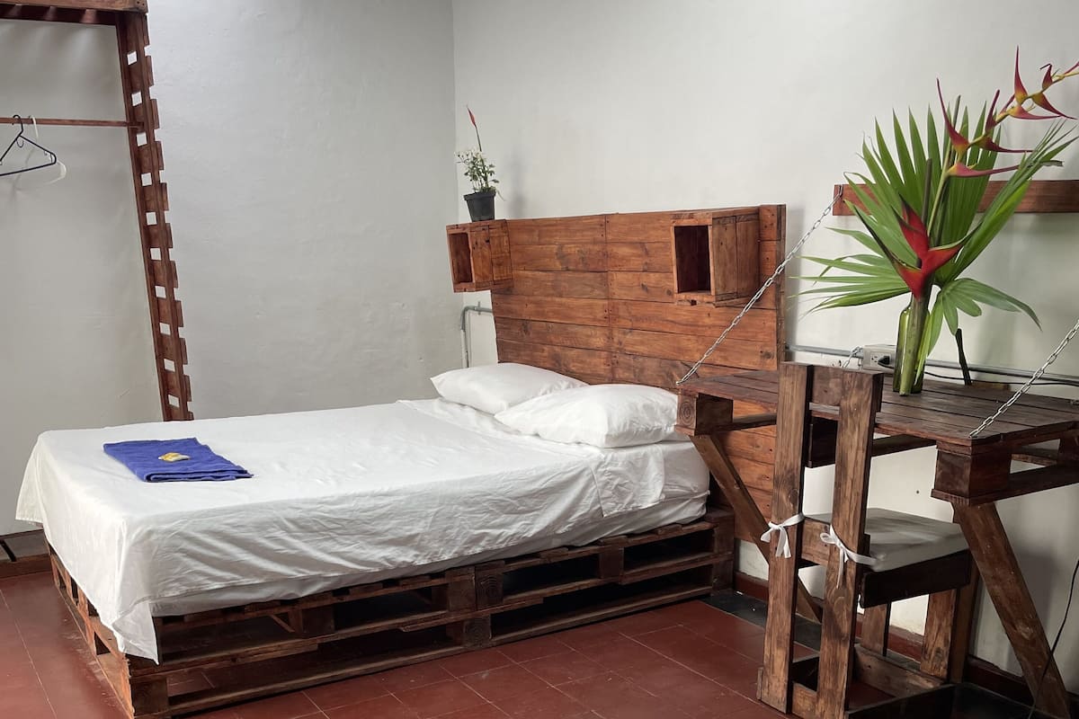Une chambre avec un lit en bois, du linge de lit blanc, un bureau en bois orné d’une plante et un luminaire suspendu.