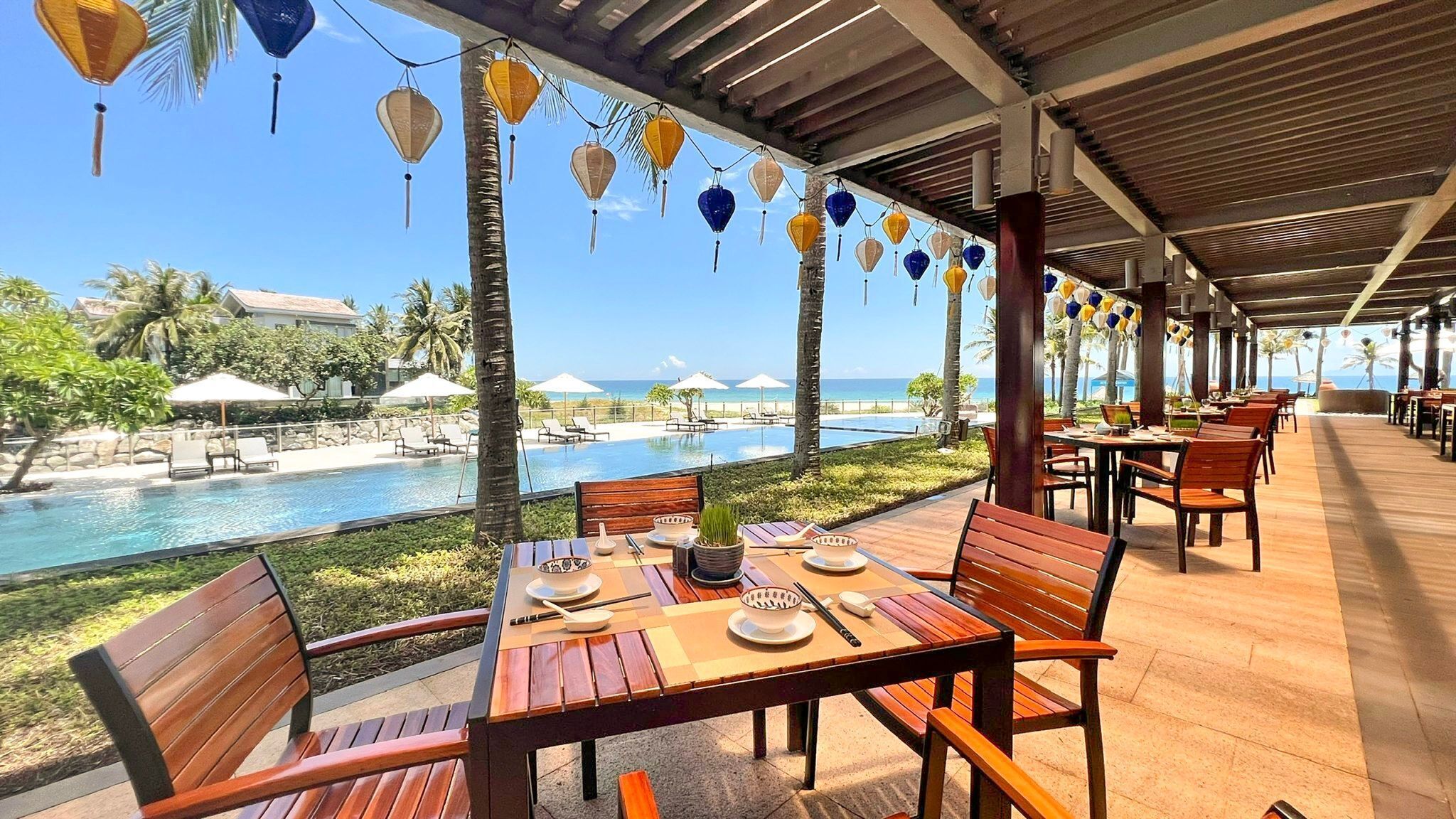 Ein Essbereich im Freien mit Holztischen und -stühlen, mit Blick auf einen Pool und einen Strand.