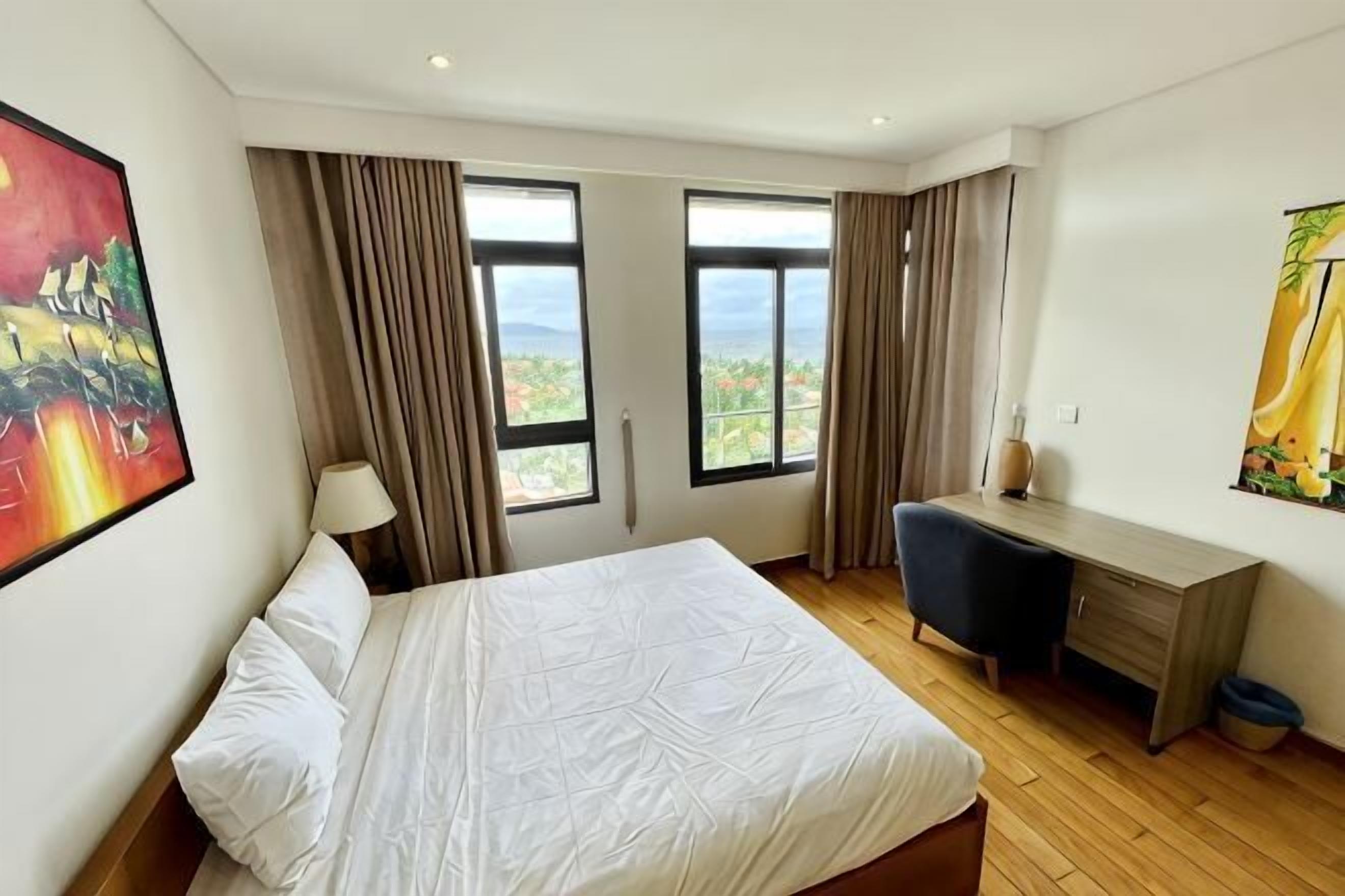 A hotel room with a bed, desk, and large windows with curtains.