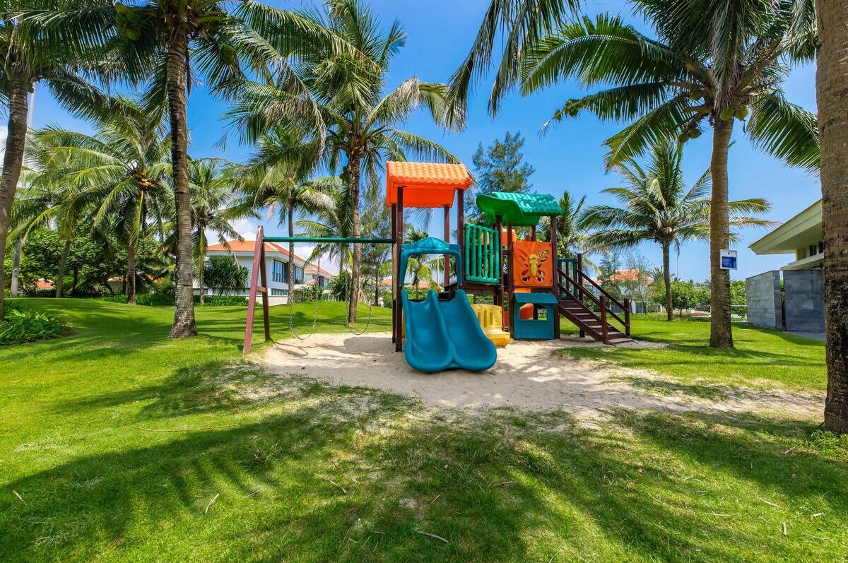 A playground with a slide and climbing structures, surrounded by palm trees and a grassy area.